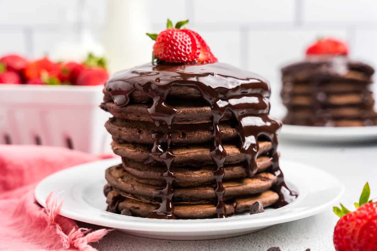 Chocolate Pancakes stacked on plate dripping with chocolate sauce topped with a fresh sliced strawberry