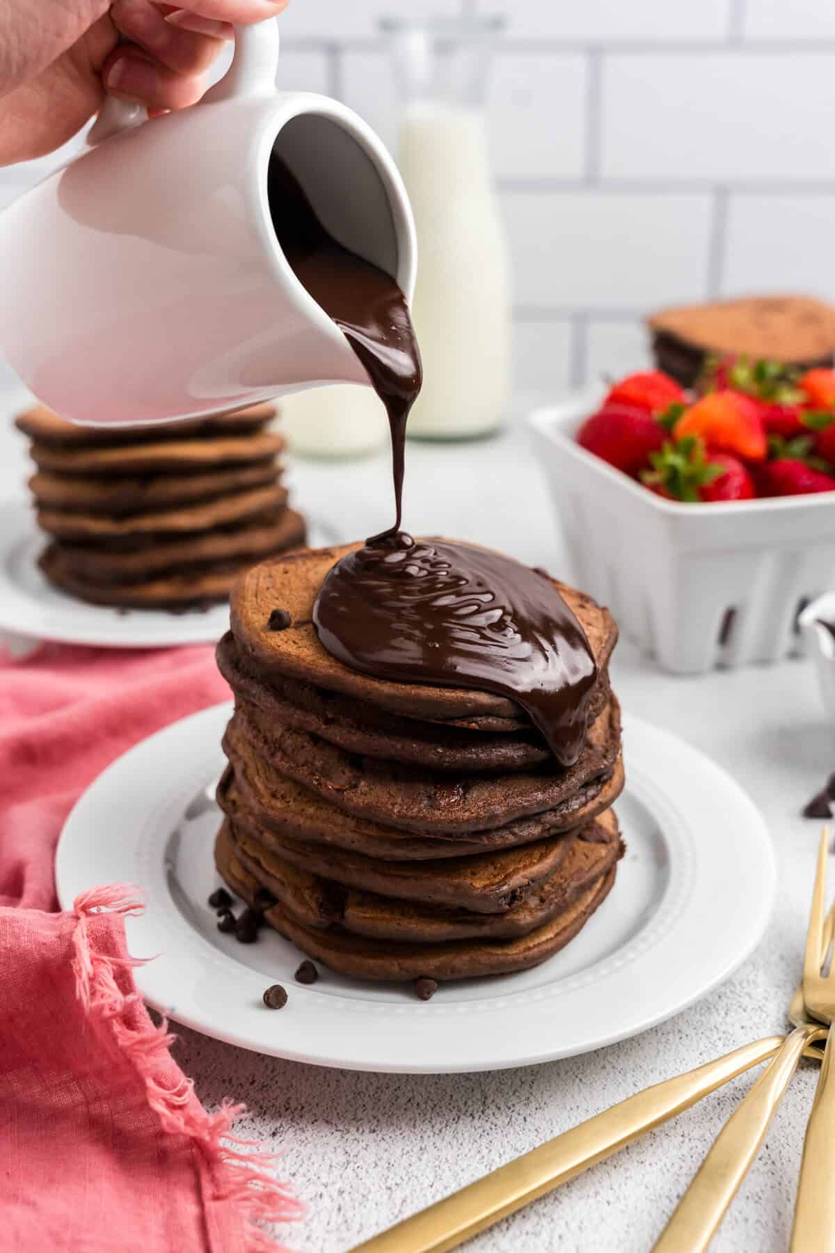 Chocolate Pancakes stacked on white plate with chocolate sauce poured overtop