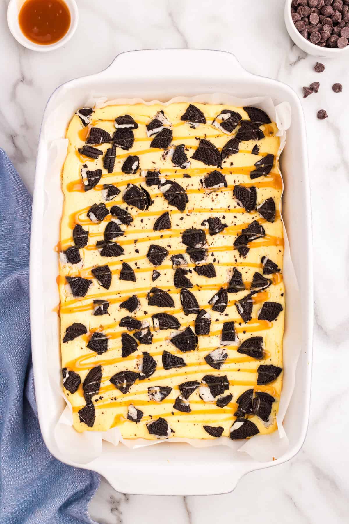 Drizzling caramel over baked Slutty Cheesecake Bars in parchment lined pan