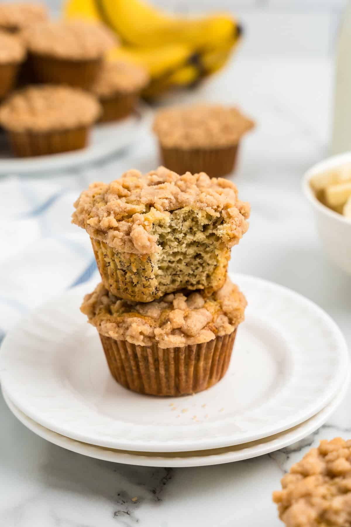 Banana Crumb Muffins stacked on white plate with one bite taken of the top muffin