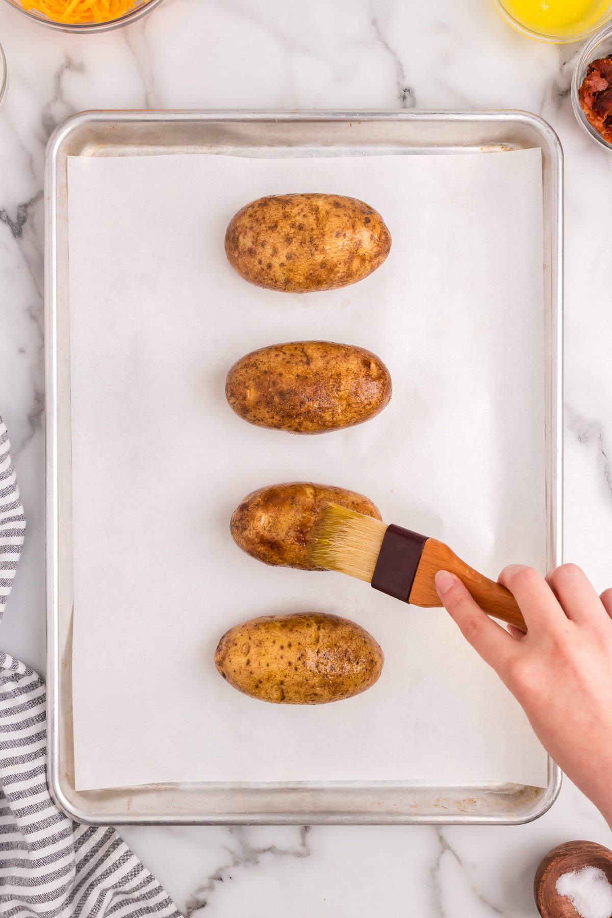 Using brush to wipe on olive oil to russet potatoes on parchment-lined baking sheet for the Twice Baked Potatoes recipe