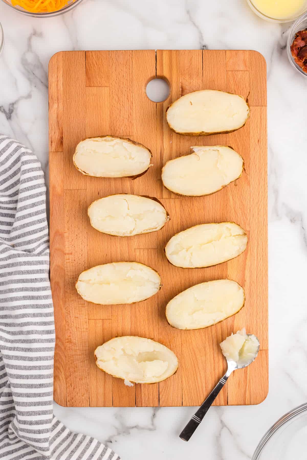 Slicing open the baked potatoes on wooden cutting board and using spoon to remove the potato from the skin for Twice Baked Potatoes recipe