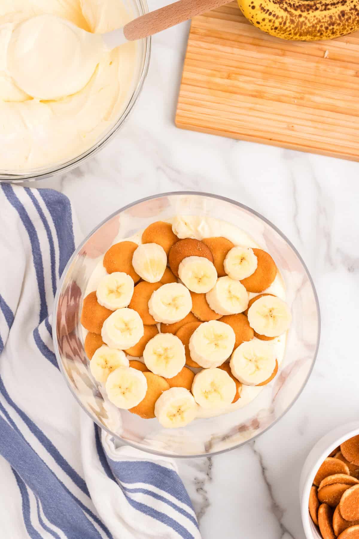 Layering Banana Pudding with vanilla wafers and sliced fresh bananas in glass footed serving bowl