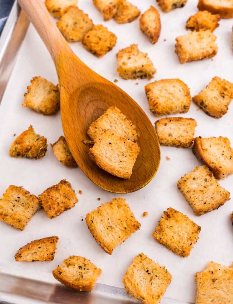 Using a wooden spoon to scoop Homemade Croutons on parchment-lined baking sheet