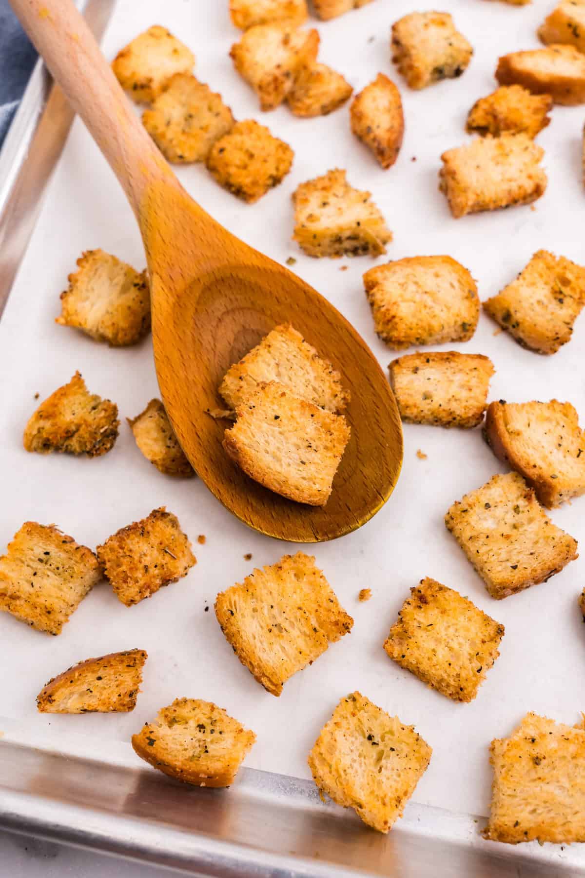 Using a wooden spoon to scoop Homemade Croutons on parchment-lined baking sheet