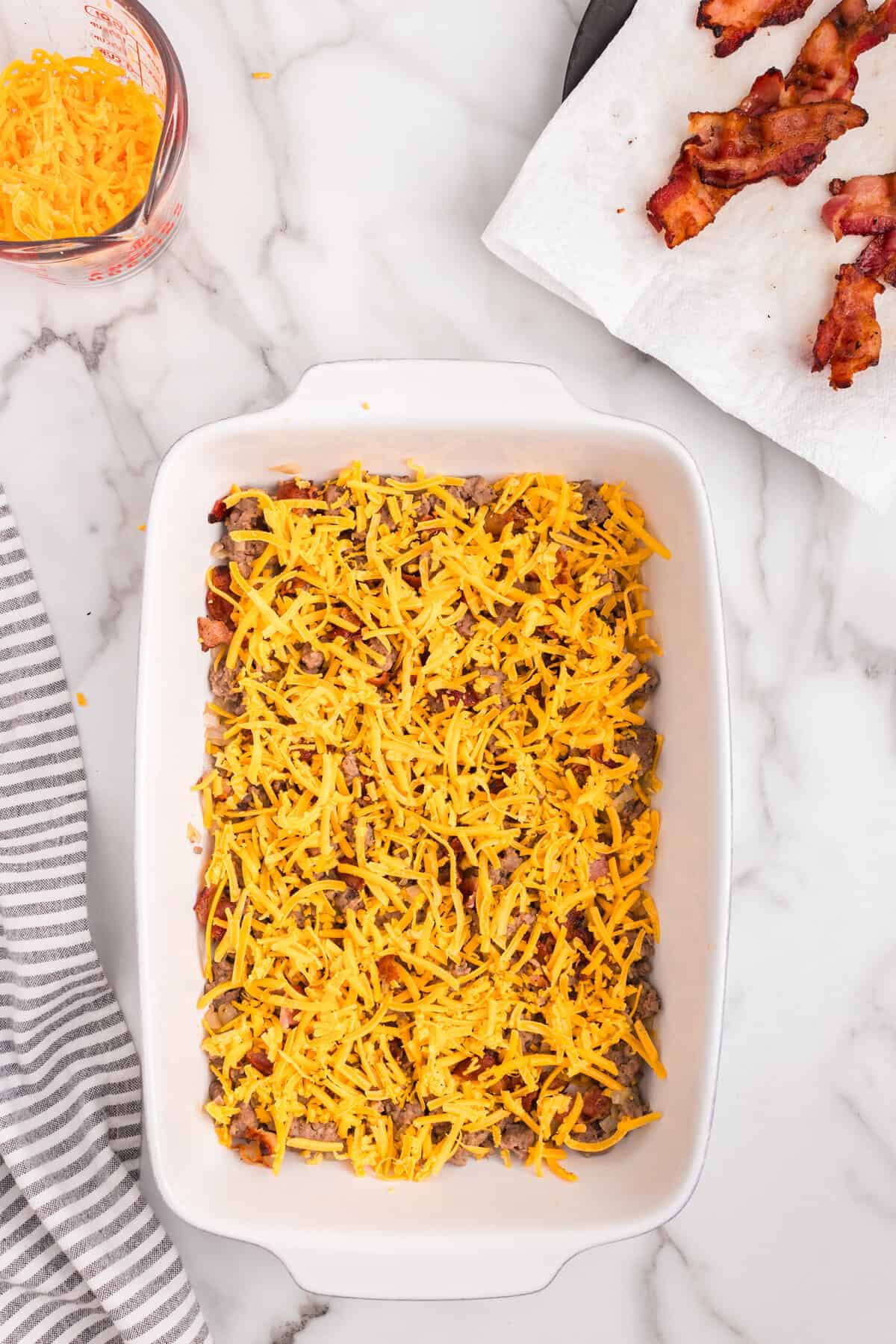 Overhead picture of cheese layered on cooked sausage and bacon in baking pan.