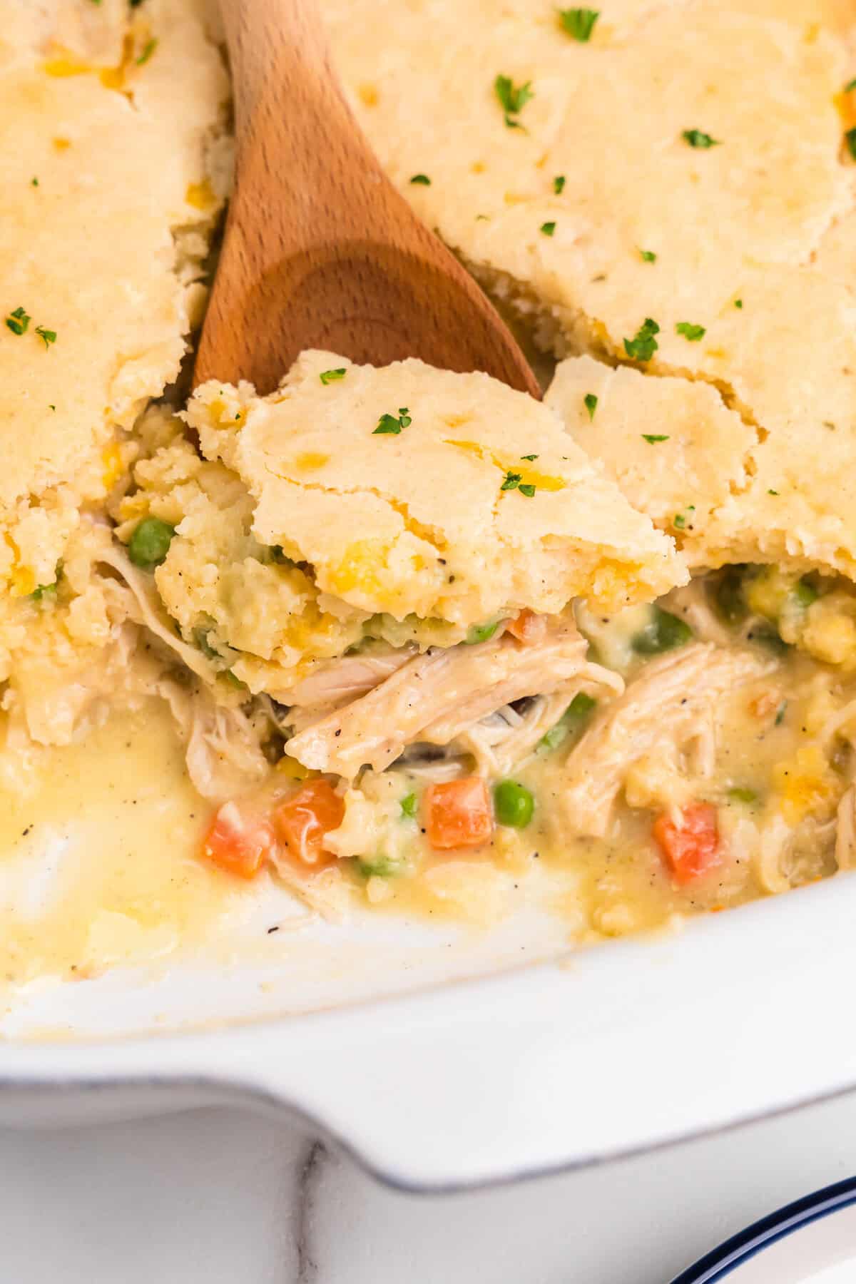 Side profile picture of white pan with a wooden spoon scooping chicken cobbler out of it.