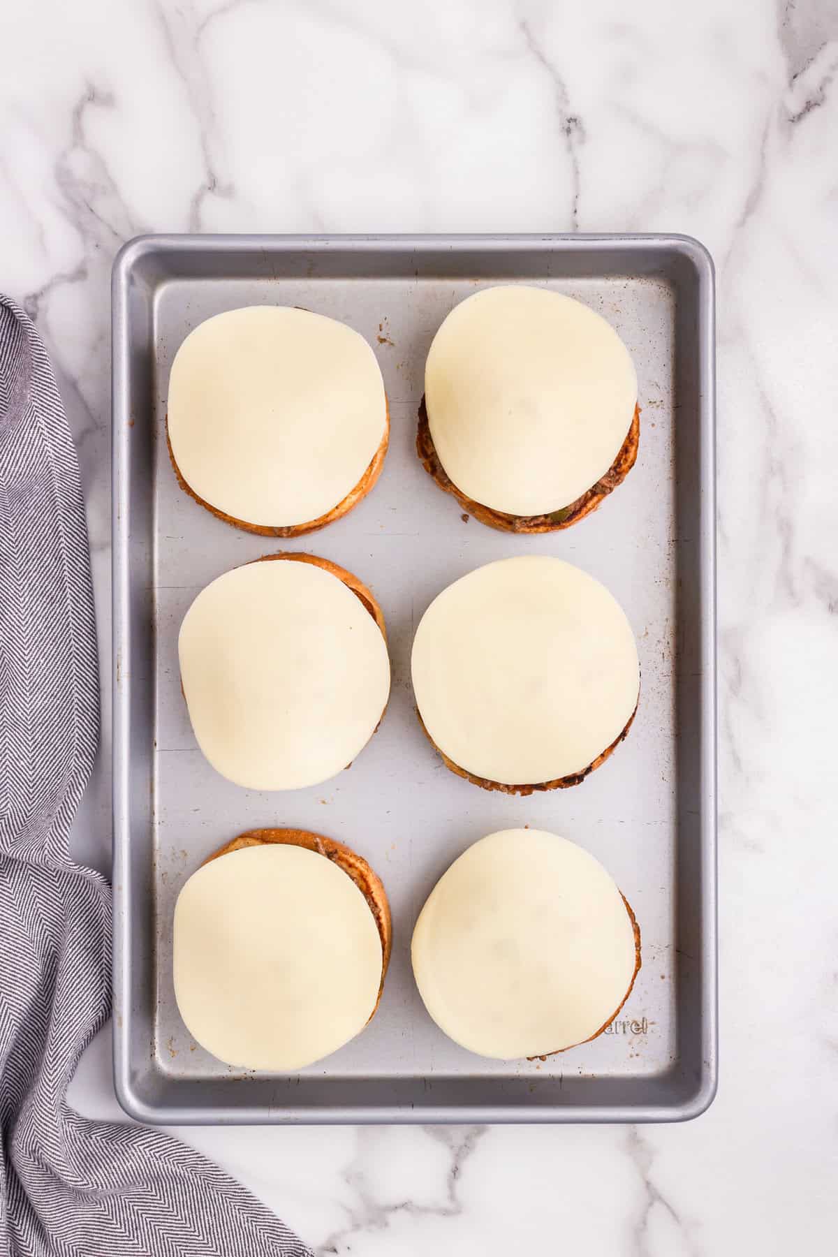 Overhead picture of metal baking sheet with 6 buns with meat and provolone cheese on them uncooked.