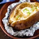 A baked potato served in a brown bowl, wrapped partially in aluminum foil. The potato is split open with a pat of butter melting on top, garnished with chopped green herbs.
