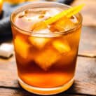 A close-up of an Old Fashioned cocktail served in a short glass with large ice cubes, garnished with an orange twist. The amber-colored drink is placed on a rustic wooden surface.