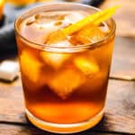 A close-up of an Old Fashioned cocktail served in a short glass with large ice cubes, garnished with an orange twist. The amber-colored drink is placed on a rustic wooden surface.