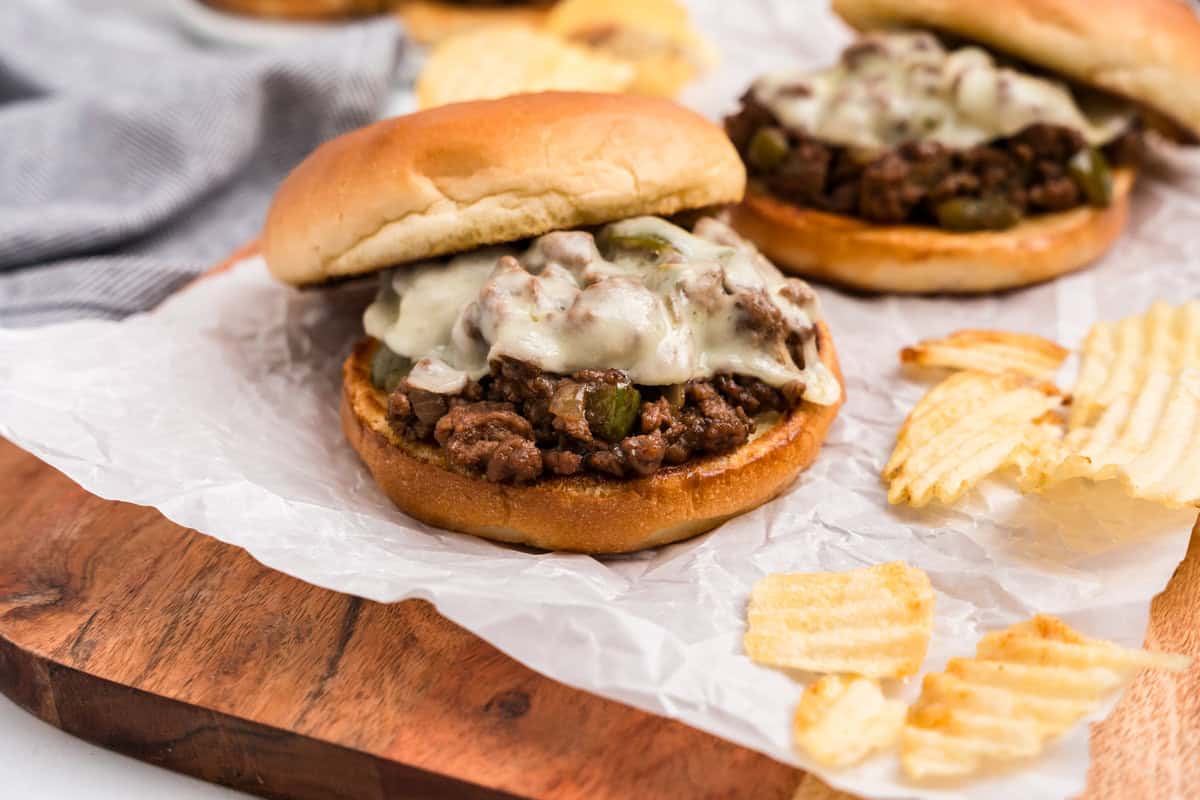 Two Cheesesteak Sloppy Joes and potato chips on a wooden cutting board.