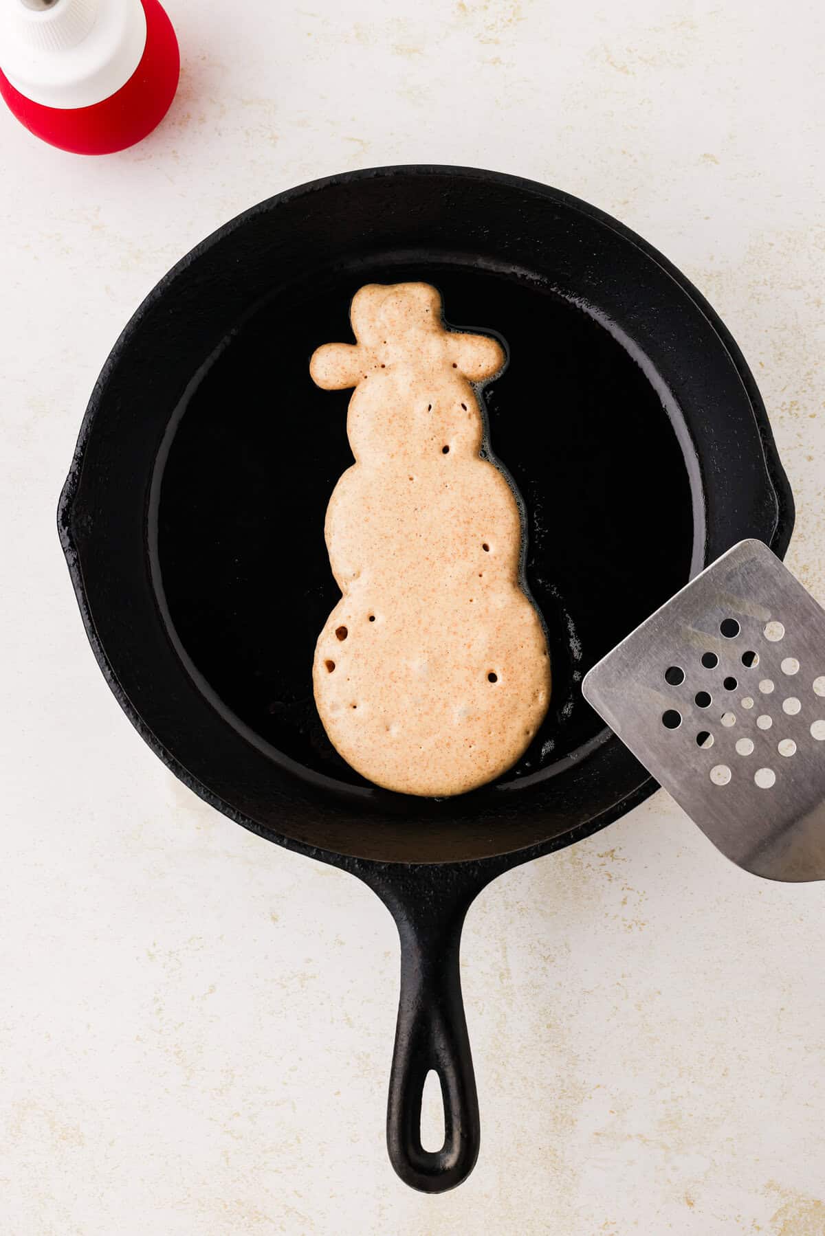 Snowman pancake cooked on one side on a skillet with a metal spatula next to it.