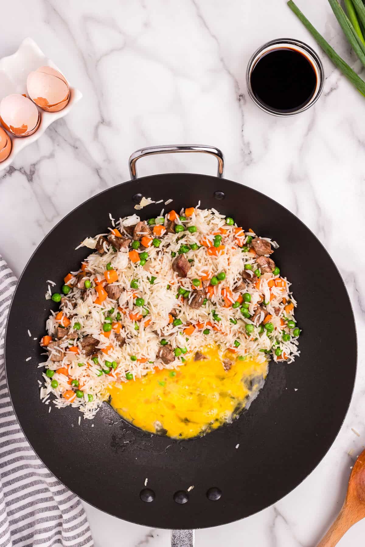 Overhead picture of black skillet pan with rice, carrots, peas and steak pushed to the side with two cracked eggs in the middle.
