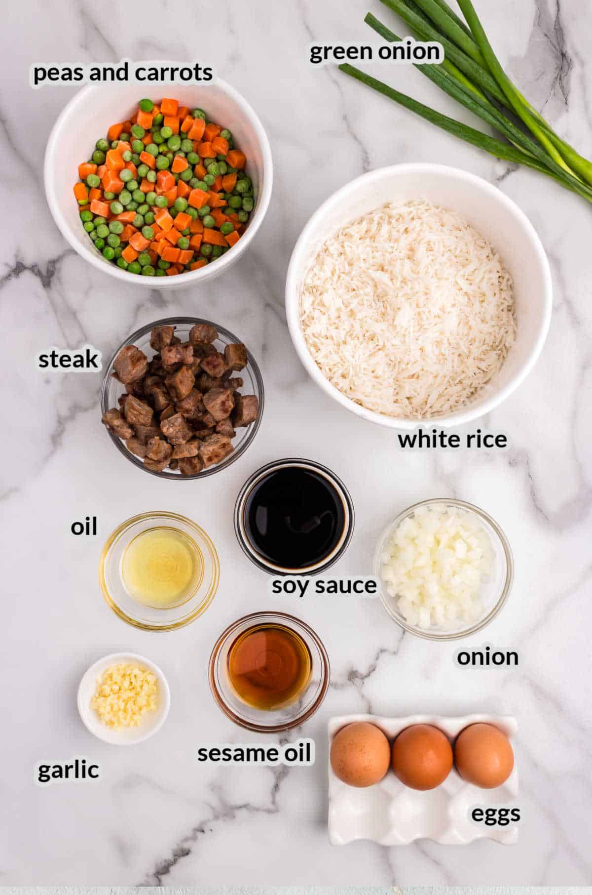 Overhead view of ingredients for steak fried rice, arranged on a marble countertop. Includes bowls of white rice, diced steak, peas and carrots, chopped onion, soy sauce, sesame oil, regular oil, and minced garlic. Three brown eggs in a carton and green onions are also present. Each ingredient is labeled with black text.