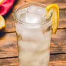 A tall glass filled with ice and a clear, bubbly liquid, garnished with a lemon wedge on the rim. The drink is placed on a rustic wooden table with a blurred background featuring a red cloth and additional lemon slices.