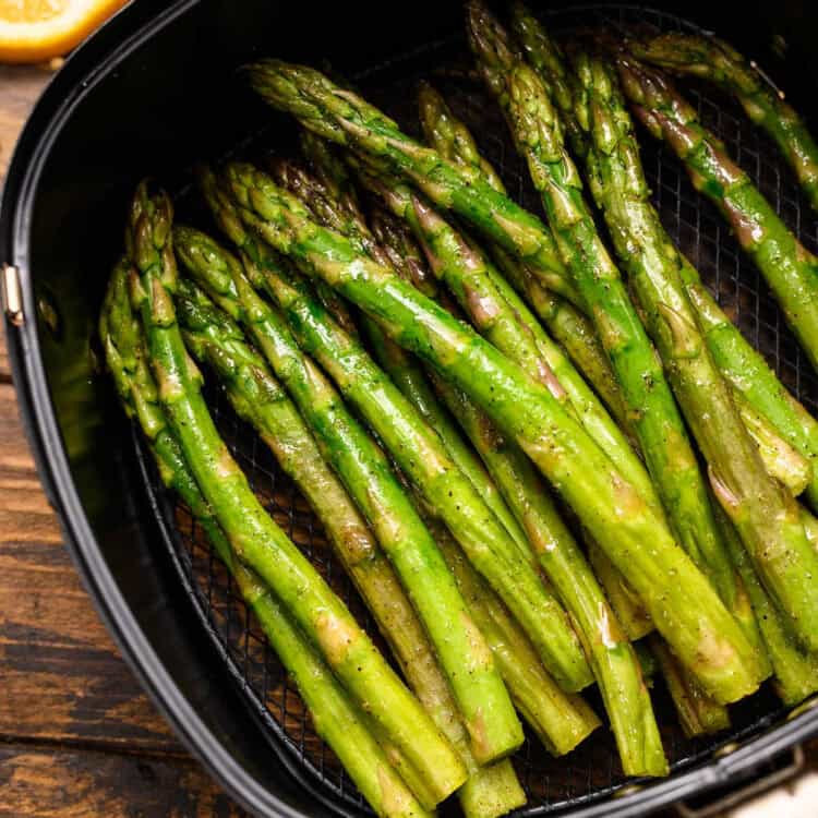Close-up of bright green asparagus spears cooked in an air fryer, lightly seasoned and glistening with oil. The asparagus is arranged in a black air fryer basket with a mesh bottom, placed on a rustic wooden surface. A lemon wedge is partially visible in the background.