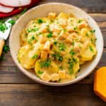 A bowl of creamy scalloped potatoes with diced ham, garnished with freshly chopped parsley. The dish is served in a rustic, off-white ceramic bowl on a wooden table, with a knife and slices of ham visible in the background. The creamy sauce coats the potatoes and ham, giving the dish a rich and comforting appearance.