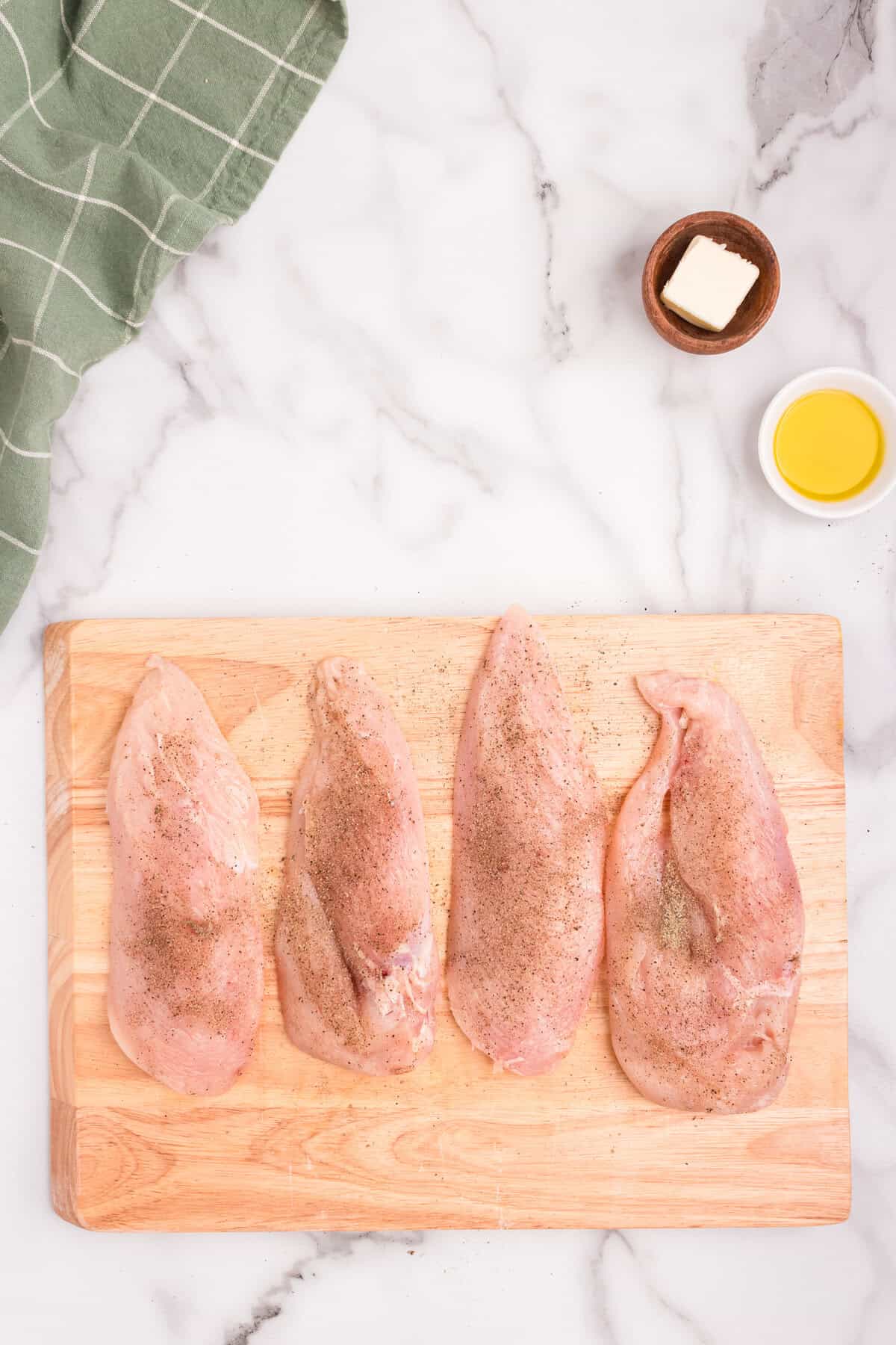 Overhead picture of raw chicken breast on a wooden cutting board with oil and butter in the corner of the picture.