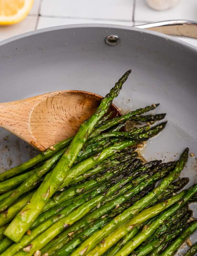 Close up image of pan fried asparagus in a grey pan with a wooden spoon scooping them.
