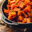 This image showcases a dish of roasted sweet potatoes, cut into cubes and seasoned with spices. The sweet potatoes have a caramelized exterior and are garnished with chopped chives. They are served in a rustic black dish on a wooden table, creating a warm and inviting presentation.