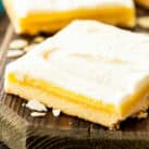 A close-up of an almond bar with a buttery shortbread crust, a smooth almond-flavored filling, and a creamy frosting on top. The bar is placed on a rustic wooden surface with almond slices scattered around.