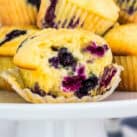 A close-up of a freshly baked blueberry muffin with a golden, fluffy texture and juicy blueberries bursting inside. The muffin is partially unwrapped, revealing its soft interior, and is placed on a white cake stand with more muffins in the background.