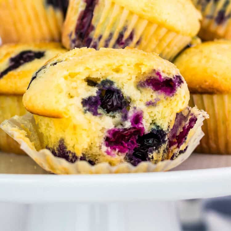 A close-up of a freshly baked blueberry muffin with a golden, fluffy texture and juicy blueberries bursting inside. The muffin is partially unwrapped, revealing its soft interior, and is placed on a white cake stand with more muffins in the background.