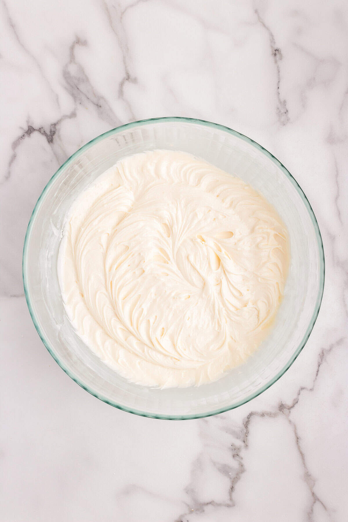 A glass mixing bowl on a marble countertop filled with smooth, creamy frosting, likely cream cheese frosting. The frosting has a swirled texture on the surface.