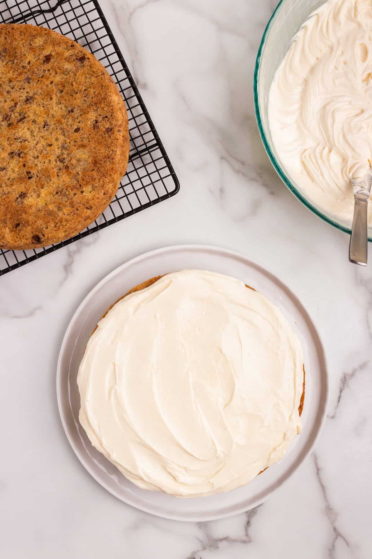 A partially assembled hummingbird cake with one cake layer on a white plate, covered in a thick layer of cream cheese frosting. Another cake layer is cooling on a wire rack nearby, and a bowl of frosting with a spatula is visible in the background on a marble countertop.