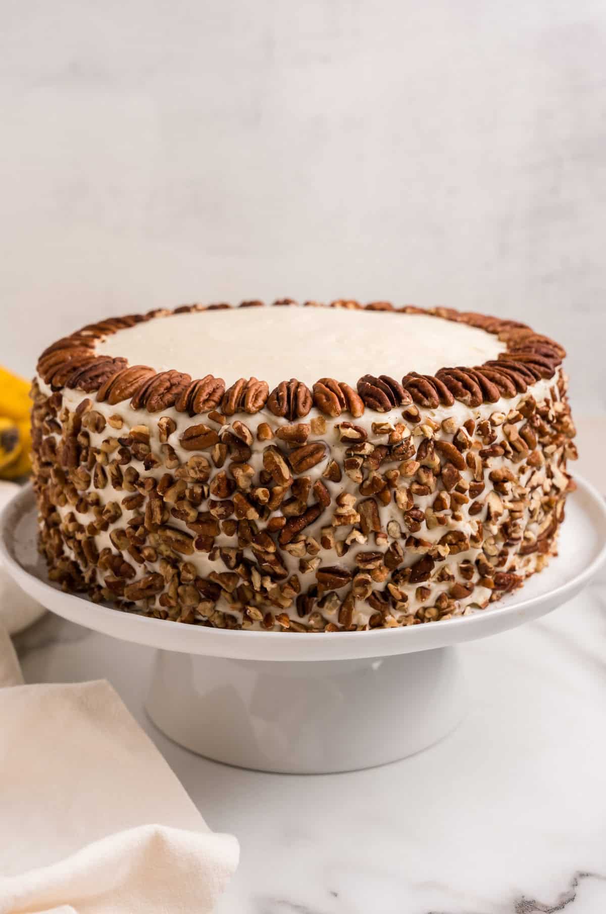 A beautifully decorated hummingbird cake on a white cake stand. The cake is covered in smooth cream cheese frosting, with chopped pecans pressed onto the sides and whole pecans neatly arranged around the top edge. A soft beige cloth napkin is draped nearby, adding to the elegant presentation.





