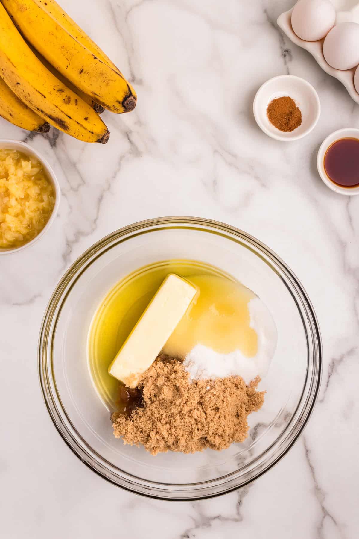 A glass mixing bowl on a marble countertop containing butter, brown sugar, white sugar, and oil. Surrounding it are ripe bananas, a bowl of crushed pineapple, a small dish of cinnamon, a small dish of vanilla extract, and a carton of eggs.