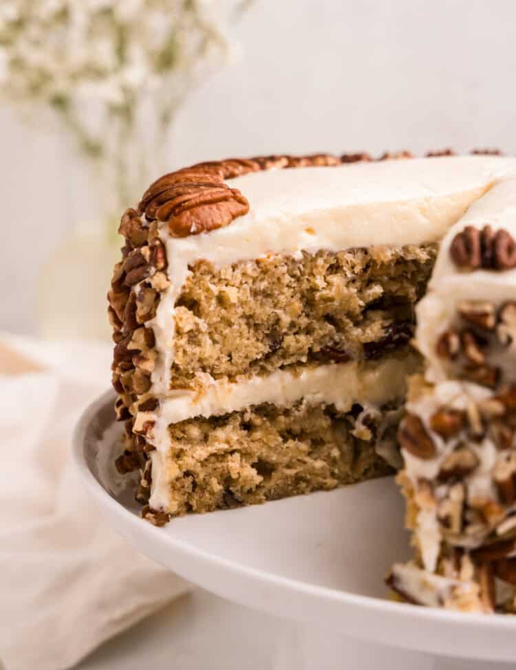 A close-up of a sliced hummingbird cake on a white cake stand, revealing its moist layers filled with bananas, pineapple, and pecans. The cake is covered in smooth cream cheese frosting, with chopped pecans pressed into the sides and whole pecans decorating the top. A blurred background with white flowers adds to the aesthetic presentation.