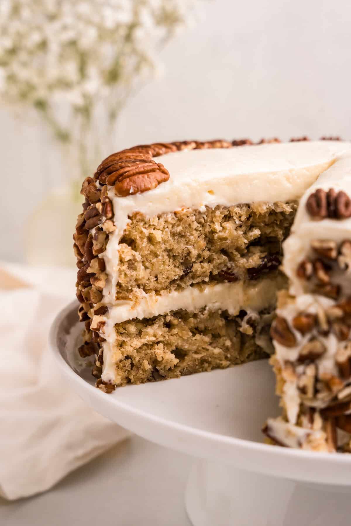A close-up of a sliced hummingbird cake on a white cake stand, revealing its moist layers filled with bananas, pineapple, and pecans. The cake is covered in smooth cream cheese frosting, with chopped pecans pressed into the sides and whole pecans decorating the top. A blurred background with white flowers adds to the aesthetic presentation.