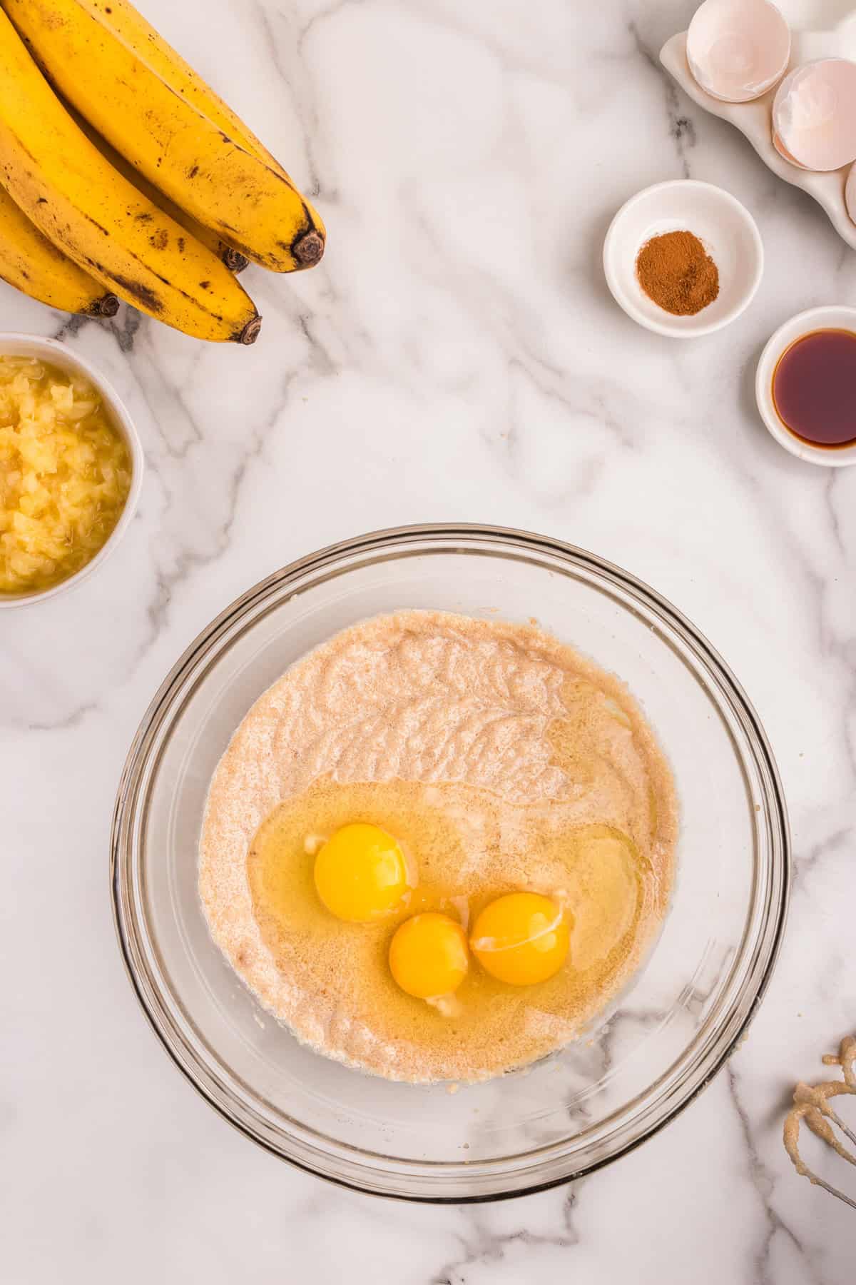A glass mixing bowl on a marble countertop containing a blended mixture of butter, sugar, and oil with three cracked eggs on top. Surrounding it are ripe bananas, a bowl of crushed pineapple, a small dish of cinnamon, a small dish of vanilla extract, and a carton of eggs with cracked shells.