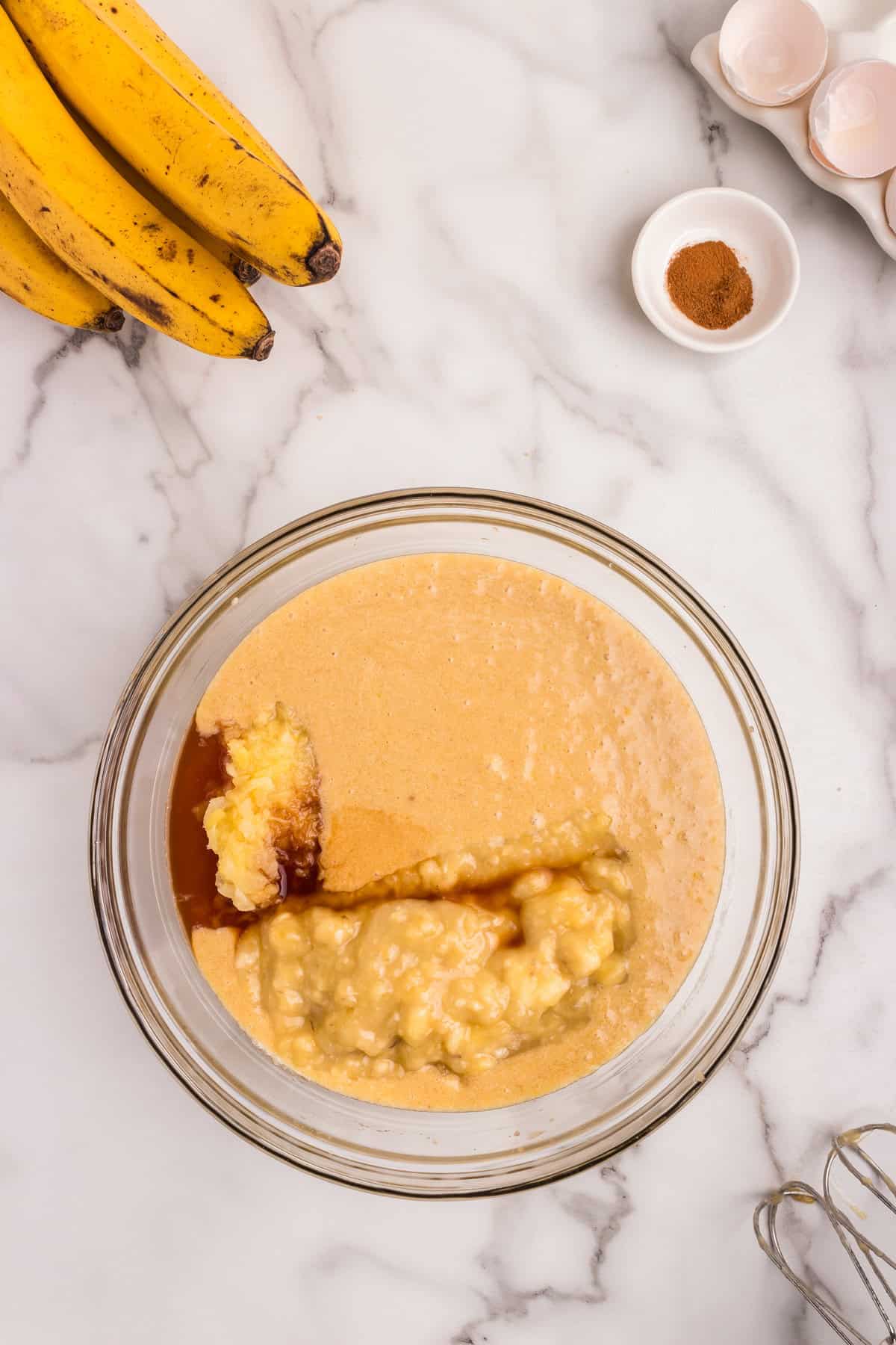 A glass mixing bowl on a marble countertop containing a blended mixture of butter, sugar, and eggs with mashed bananas, crushed pineapple, and vanilla extract added on top. Surrounding it are ripe bananas, a small dish of cinnamon, a carton of cracked eggshells, and a hand mixer.