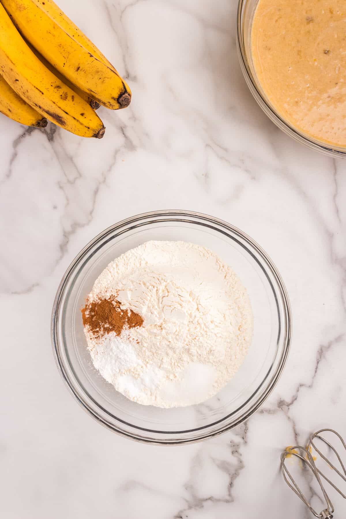 A glass mixing bowl on a marble countertop containing dry ingredients, including flour, cinnamon, baking soda, and sugar. Surrounding it are ripe bananas, a separate bowl with the wet mixture, and a hand mixer with batter-coated beaters.