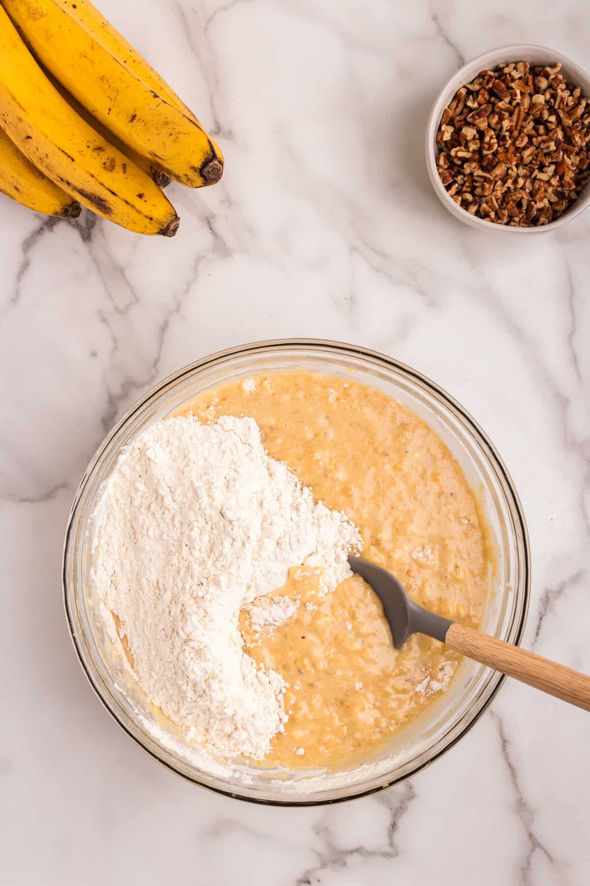A glass mixing bowl on a marble countertop containing a wet cake batter mixture with a portion of dry ingredients being folded in using a spatula. Surrounding it are ripe bananas and a bowl of chopped pecans.