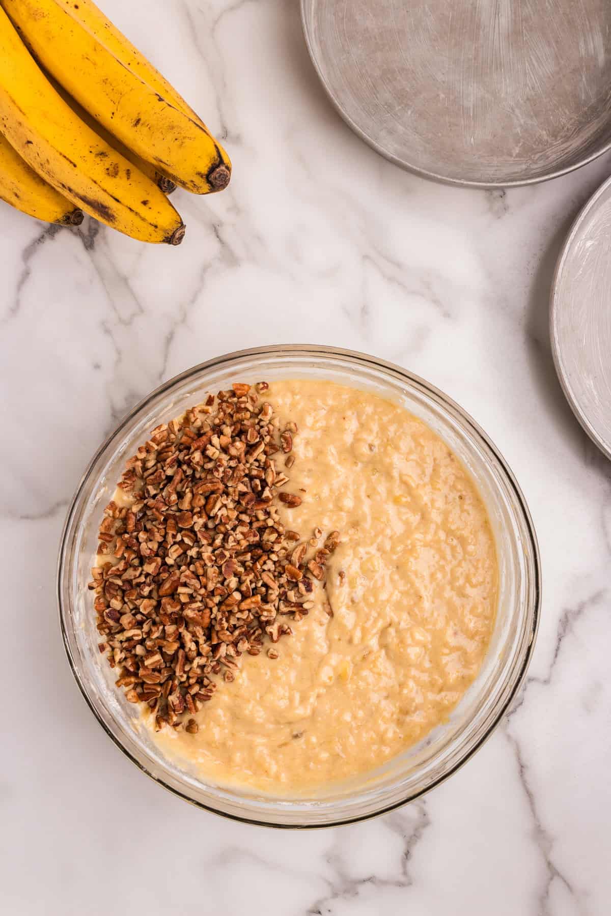 A glass mixing bowl on a marble countertop containing hummingbird cake batter with chopped pecans added on top. Surrounding it are ripe bananas and greased cake pans ready for baking.