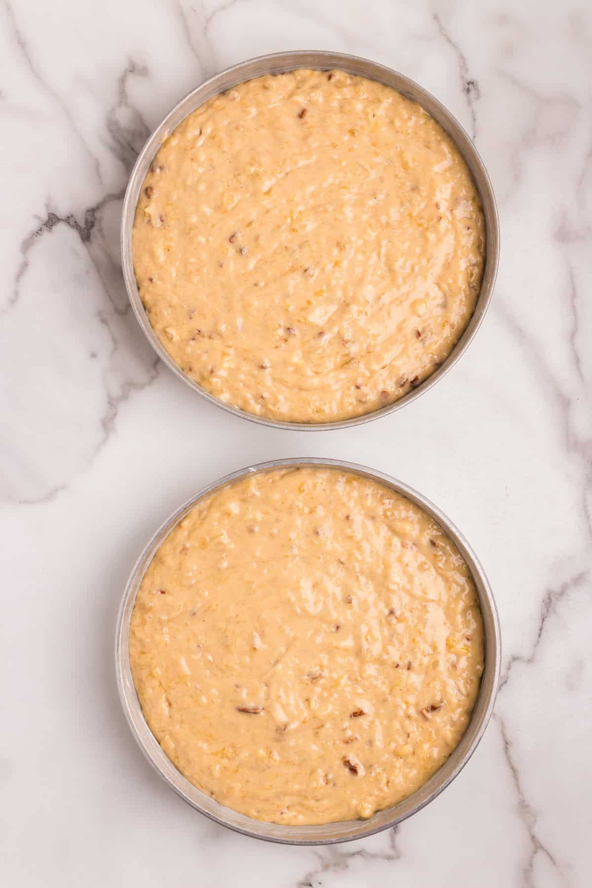 Two round cake pans filled with hummingbird cake batter, ready for baking. The batter contains visible pieces of chopped pecans, mashed bananas, and crushed pineapple. The pans are placed on a marble countertop.
