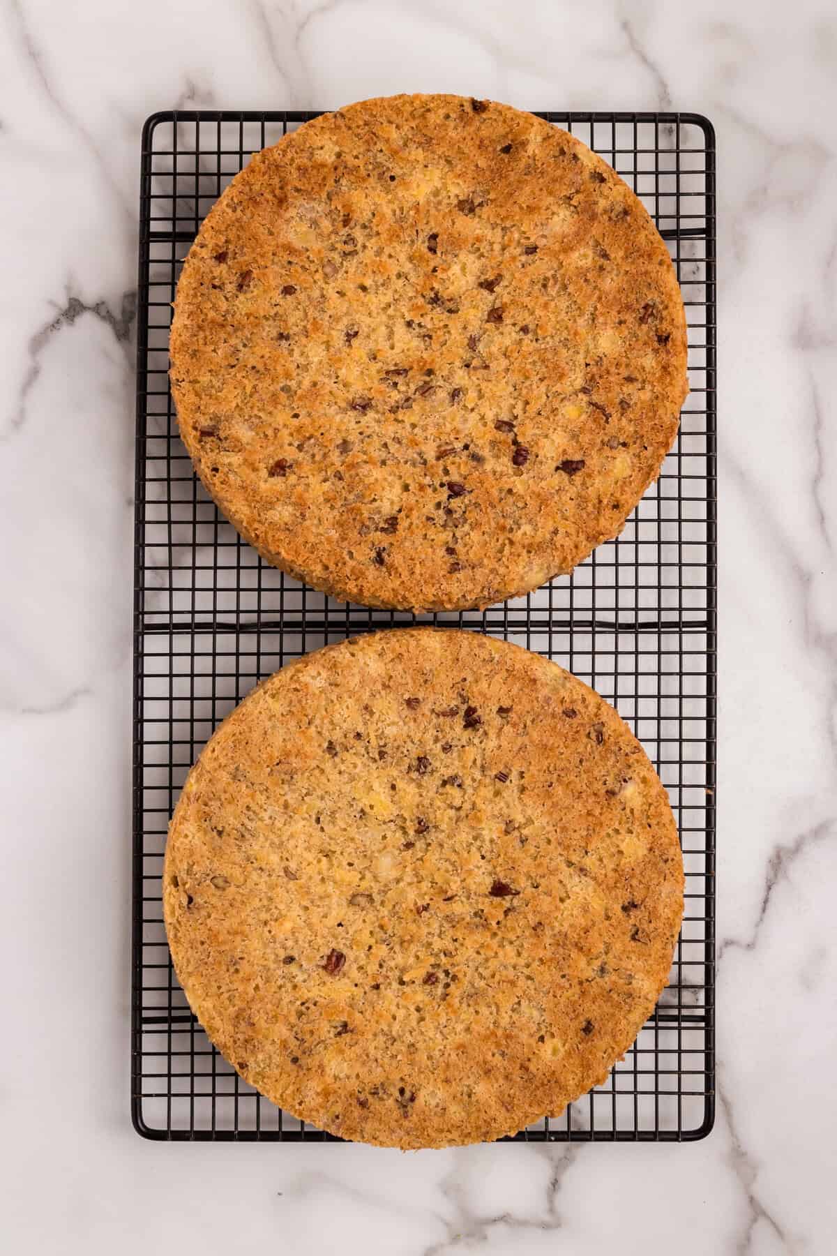 Two freshly baked hummingbird cake layers cooling on a black wire rack. The cakes have a golden-brown color with visible bits of pecans, bananas, and pineapple. The rack is placed on a marble countertop.