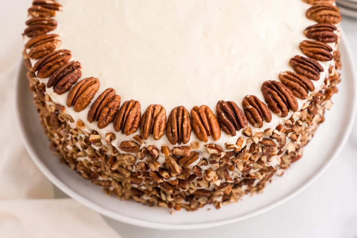 A beautifully decorated hummingbird cake covered in smooth cream cheese frosting. The sides are coated with chopped pecans, and whole pecans are arranged neatly around the top edge. The cake sits on a white plate, creating an elegant presentation.