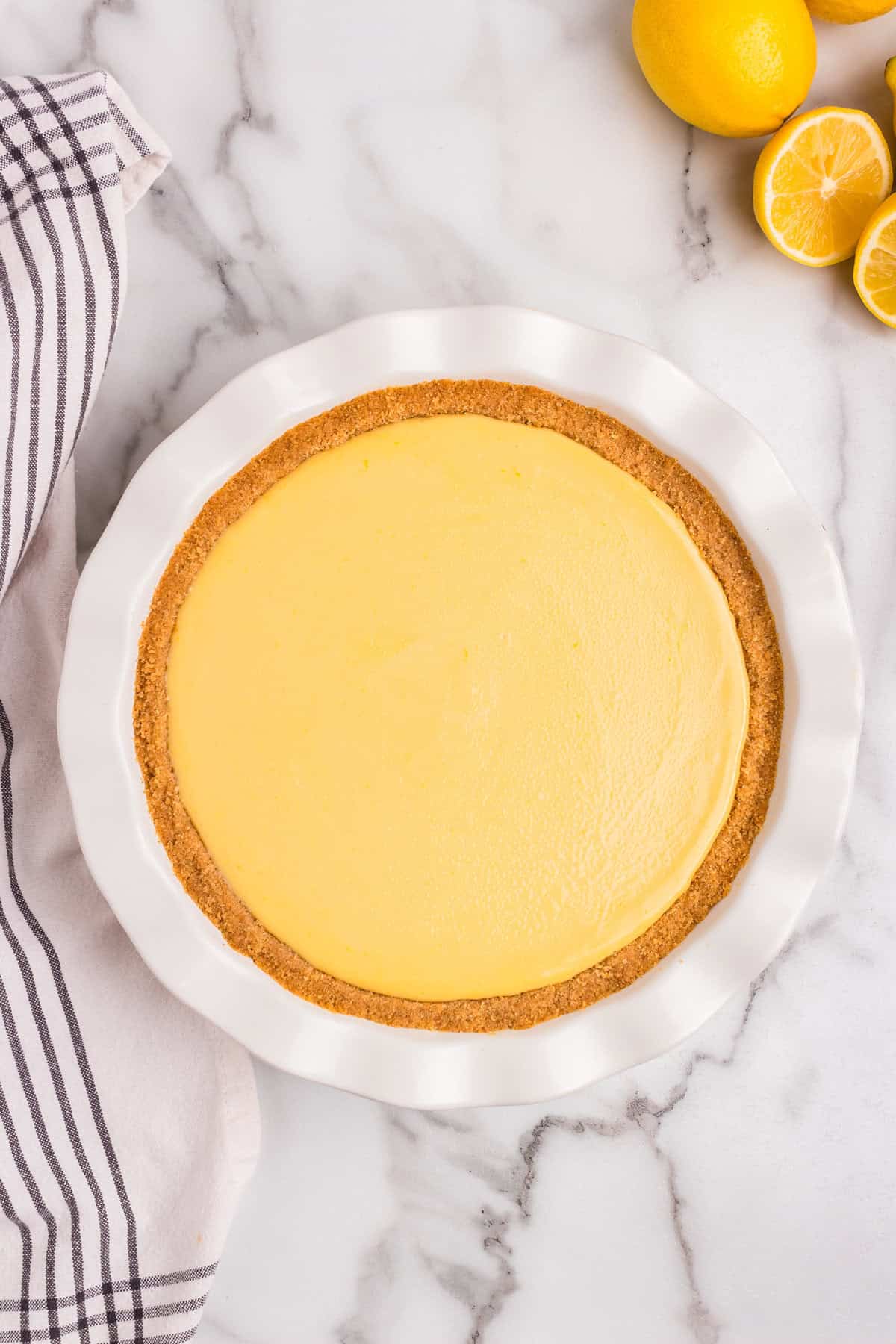 A freshly baked lemon pie with a smooth, golden-yellow filling sits in a white ceramic pie dish. The crust is golden brown and evenly baked. The pie is placed on a marble countertop with a striped kitchen towel beside it. Fresh lemons, some whole and some sliced, are arranged nearby.