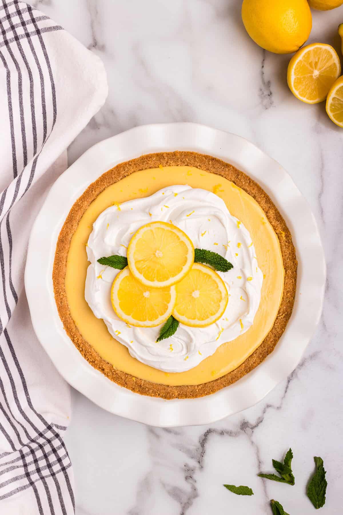 A beautifully decorated lemon pie with a golden graham cracker crust, topped with swirls of whipped cream, fresh lemon slices, and mint leaves. Bright yellow lemon zest is sprinkled on top, enhancing the fresh citrus look. The pie sits in a white ceramic dish on a marble countertop, with whole and halved lemons nearby. A striped kitchen towel and scattered mint leaves add a cozy, inviting touch to the scene.