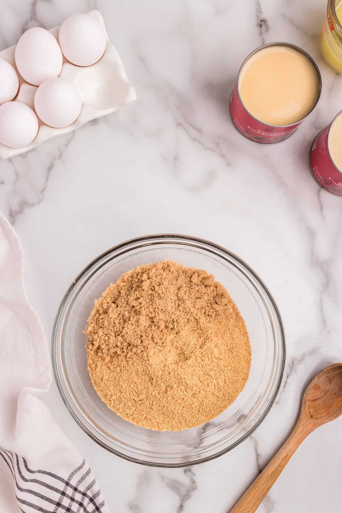 A glass mixing bowl filled with finely ground graham cracker crumbs sits on a marble countertop. Nearby, a wooden spoon rests on the surface. In the background, a carton of eggs, open cans of sweetened condensed milk, and a measuring cup are visible. A white and striped kitchen towel is partially draped on the left side.