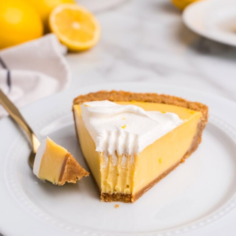 A close-up of a slice of lemon pie on a white plate, with a bite taken out and resting on a gold fork. The pie has a creamy, smooth lemon filling with a golden graham cracker crust, topped with fluffy whipped cream and a sprinkle of lemon zest. In the blurred background, the remaining pie is visible in a white ceramic dish, along with fresh whole and halved lemons on a marble countertop.