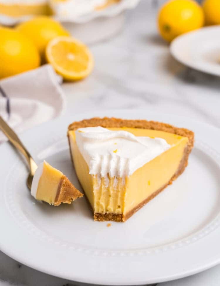A close-up of a slice of lemon pie on a white plate, with a bite taken out and resting on a gold fork. The pie has a creamy, smooth lemon filling with a golden graham cracker crust, topped with fluffy whipped cream and a sprinkle of lemon zest. In the blurred background, the remaining pie is visible in a white ceramic dish, along with fresh whole and halved lemons on a marble countertop.