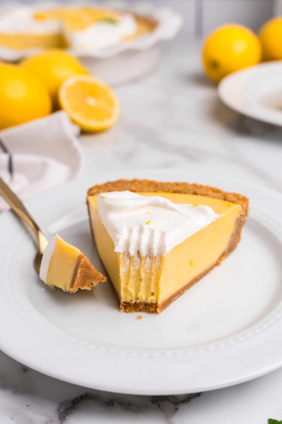 A close-up of a slice of lemon pie on a white plate, with a bite taken out and resting on a gold fork. The pie has a creamy, smooth lemon filling with a golden graham cracker crust, topped with fluffy whipped cream and a sprinkle of lemon zest. In the blurred background, the remaining pie is visible in a white ceramic dish, along with fresh whole and halved lemons on a marble countertop.