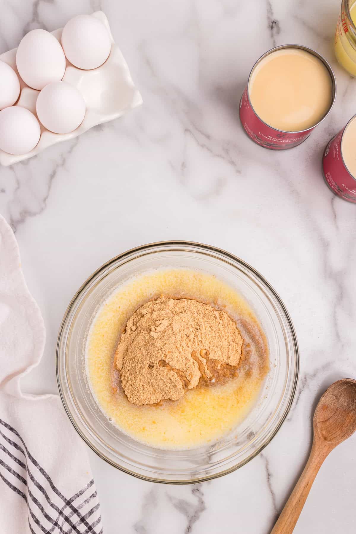A glass mixing bowl containing graham cracker crumbs and melted butter sits on a marble countertop, ready to be mixed for a pie crust. A wooden spoon is placed nearby. In the background, there is a carton of eggs, open cans of sweetened condensed milk, and a measuring cup. A white and striped kitchen towel is partially visible on the left.