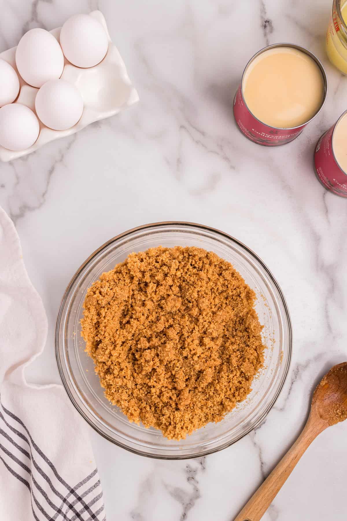 A glass mixing bowl filled with a crumbly graham cracker crust mixture sits on a marble countertop. A wooden spoon with traces of the mixture rests nearby. In the background, there is a carton of eggs, open cans of sweetened condensed milk, and a measuring cup, all ingredients for making a lemon pie. A white and striped kitchen towel is partially visible on the left.